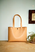 Tuscan leather tote bag with double top handles and a small sterling silver twig closure on the front. Displayed on a light wood surface against a pale blue-gray wall with a framed print and potted plant in the background.
