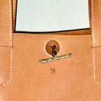 Close-up of a tan leather tote bag featuring a decorative silver-toned branch clasp with a black dog figure, a small dog logo, and white contrast stitching.