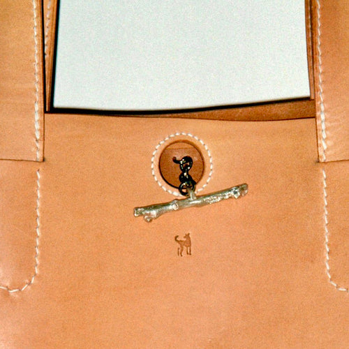 Close-up of a tan leather tote bag featuring a decorative silver-toned branch clasp with a black dog figure, a small dog logo, and white contrast stitching.